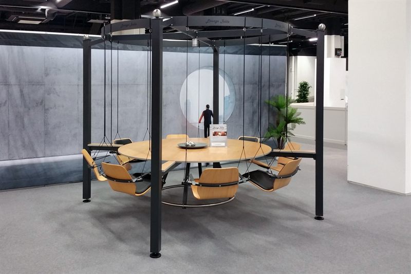 Der LOUNGETABLE freistehend: runder Holztisch im Zentrum, darum acht an Seilen hängende Holzsitze, getragen von einem dunklen Metallgestell mit vier Stützen
