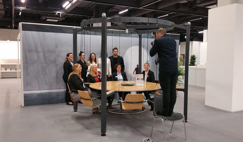 Gruppenfoto am LOUNGETABLE auf einer Messe: Gäste sitzen und stehen um den runden Holztisch mit den hängenden Schwingsitzen